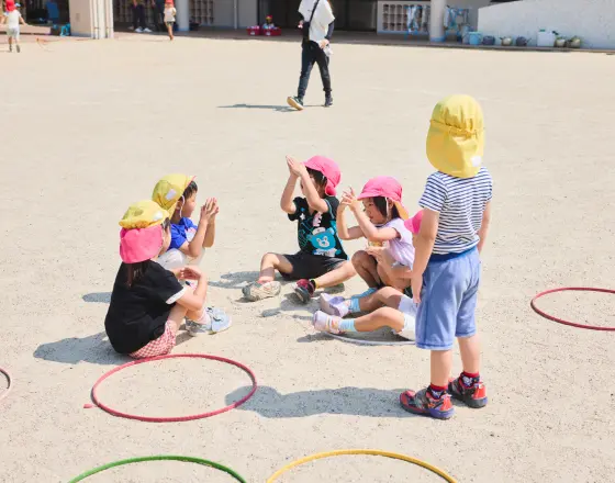 園庭で帽子をかぶった幼児たちが輪になって遊んでいる様子