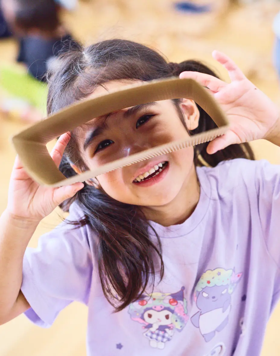 段ボールの切れ端を手に取り、笑顔で顔をのぞかせながら遊んでいる幼児の様子